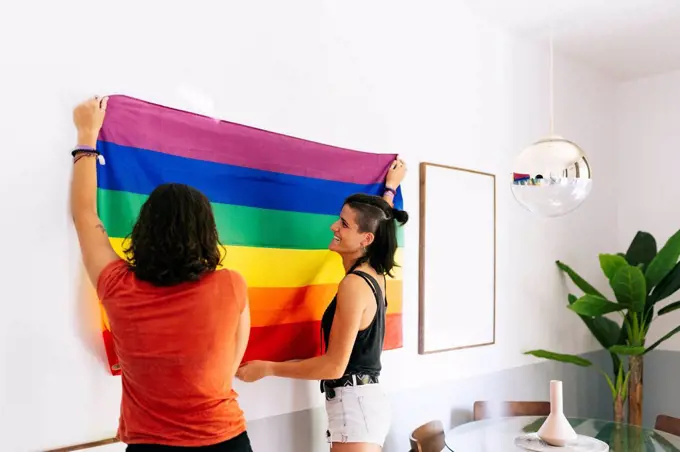 Female friends holding rainbow flag against wall at home