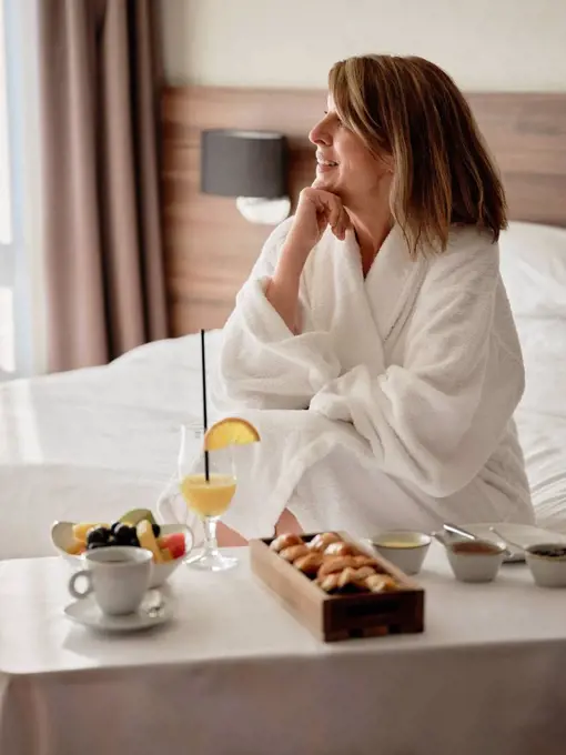 Smiling thoughtful senior woman looking away while sitting with breakfast in luxury hotel room