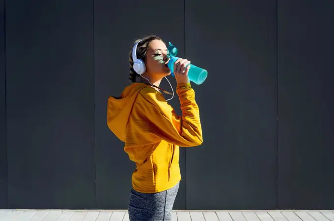 Female athlete wearing headphones drinking water while standing against gray wall