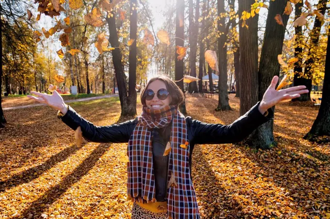 Smiling beautiful woman with arms outstretched throwing dry leaves while standing at park during autumn