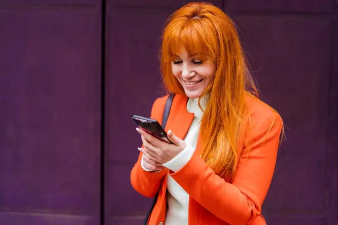 Smiling woman in jacket using mobile phone against wall