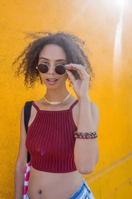 Fashionable young woman with curly hair holding sunglasses standing by yellow wall