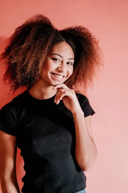 Smiling Afro woman with hand on chin standing against peach background