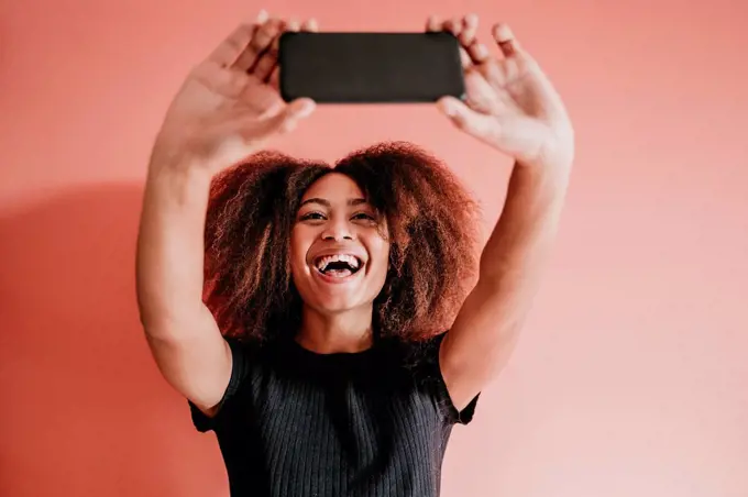 Cheerful Afro woman taking selfie through mobile phone against peach background