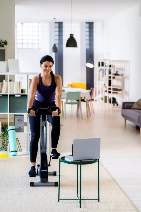 Female athlete exercising on exercise bike while using laptop at home