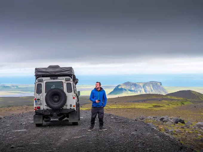 Male tourist looking away while standing by off-road vehicle on road against cloudy sky