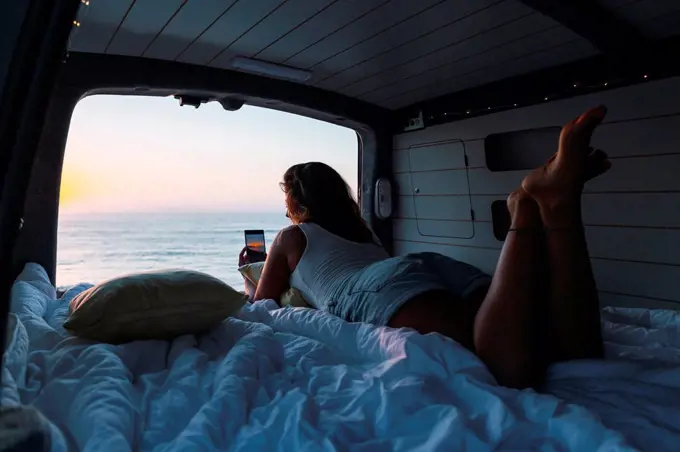 Woman taking photo of sunset through phone while lying in camper van at beach
