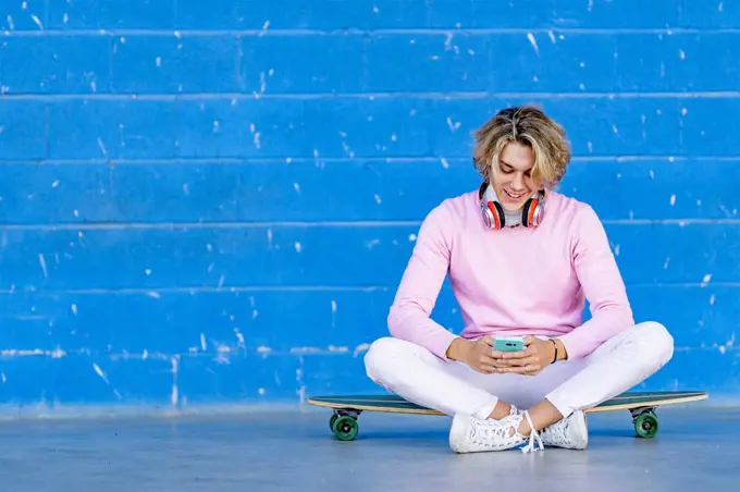 Blond man using phone while sitting on skate board against blue wall