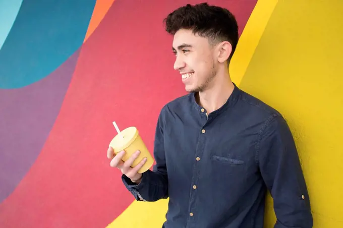 Happy young man holding disposable cup while looking away against multi colored wall