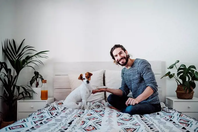 Handsome man playing with dog while sitting on bed at home