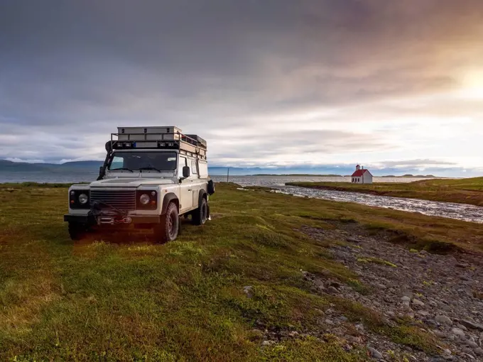 Off-road car parked in front of Icelandic river at cloudy sunset with secluded Unadsdalskirkja church in background