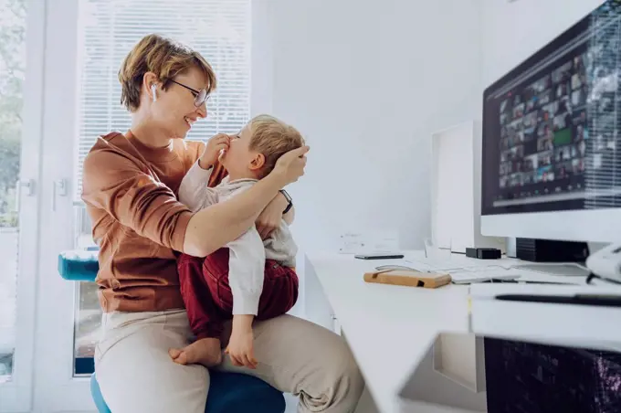 Loving mother embracing son sitting on her lap at home office