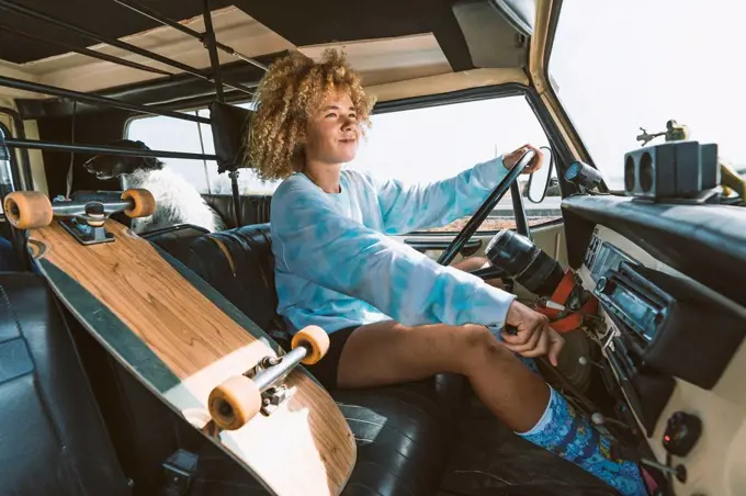 Smiling blond Afro woman sitting by skateboard while driving old off-road vehicle