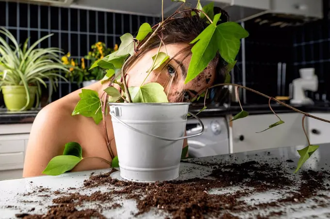 Young woman face covered with leaf and flower pot at home