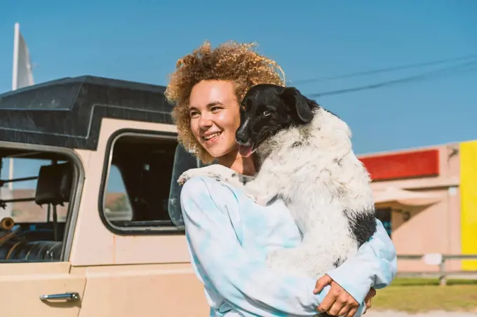 Smiling young blond woman carrying dog while looking away on sunny day