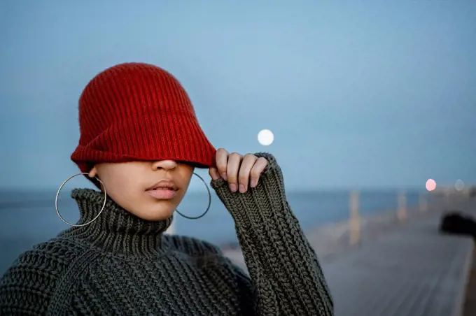 Woman covering eyes with knit hat while standing against sky