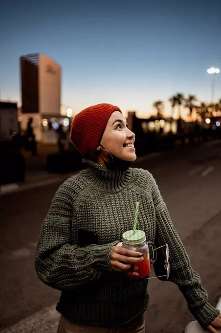 Smiling woman with face mask below chin holding juice while standing on road