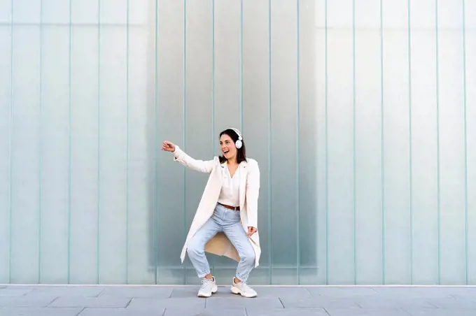 Cheerful businesswoman listening music through headphones while dancing on footpath against glass wall