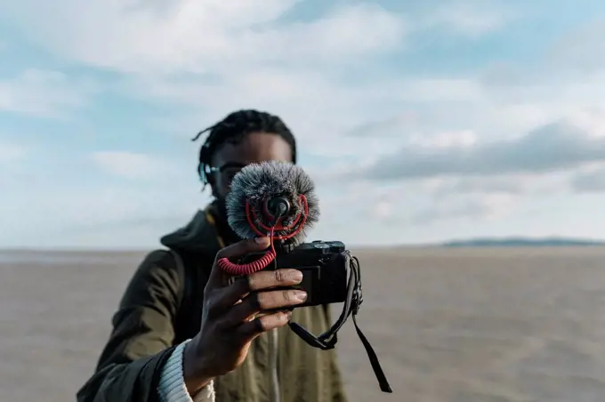 Young traveler making vlog of himself at beach against sky