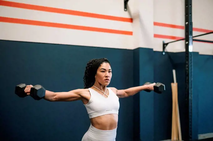 Female athlete working out with dumbbells against wall in gym