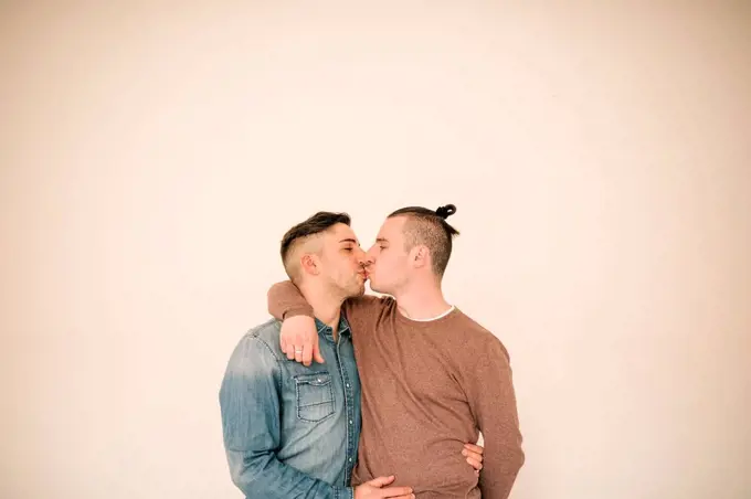 Affectionate gay couple kissing against beige background
