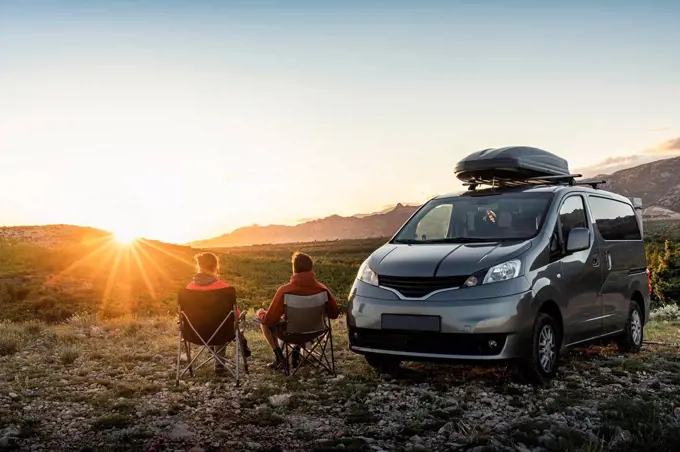 Man and woman sitting near camper van, looking at landscape at sunset