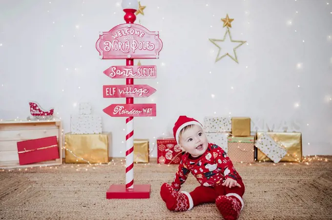 Smiling baby girl wearing Santa hat playing while sitting by direction pole at home during Christmas