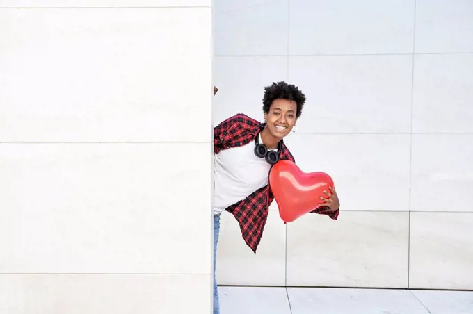 Happy young woman holding red heart shape balloon while standing behind white wall