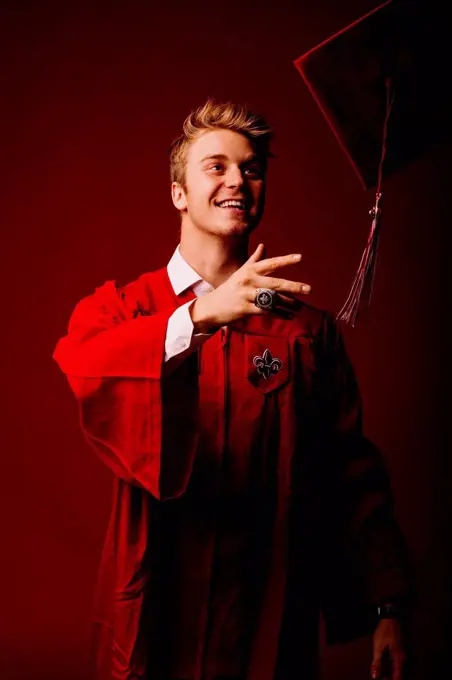 Male university student in graduation gown throwing mortarboard against red background