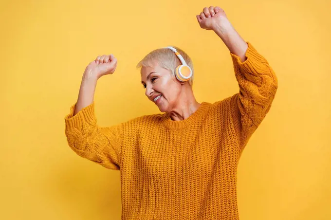 Carefree woman wearing headphones dancing with hand raised against yellow background