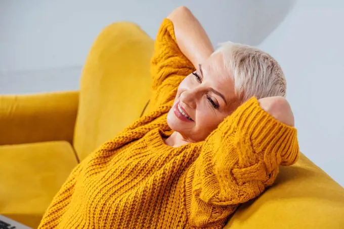 Smiling woman with hands behind back looking away while resting on sofa