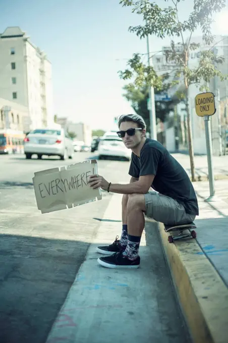 Young man holding signboard while sitting over skateboard on sidewalk