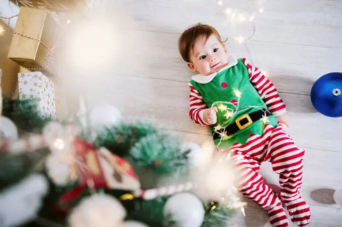 Baby boy in elf costume playing with string light while lying on floor at home during Christmas