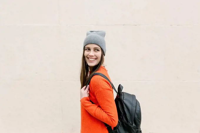 Smiling young woman with backpack looking away in front of wall