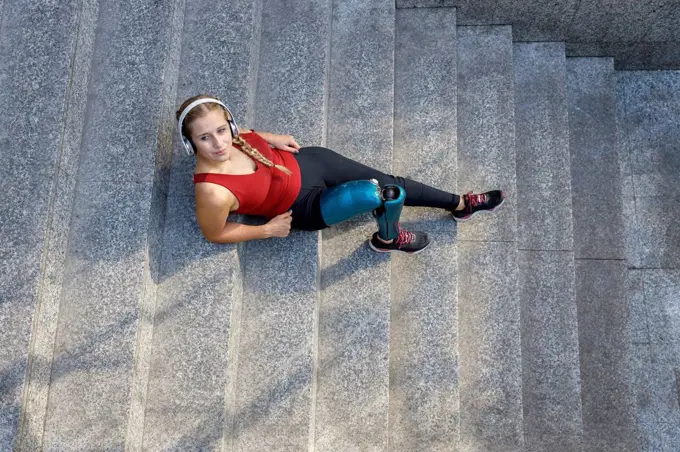 Young sportswoman with prosthetic leg relaxing while sitting on steps