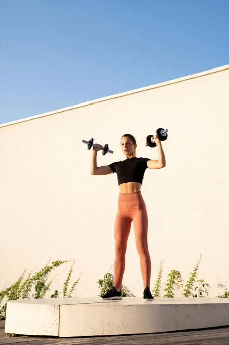 Sportswoman flexing muscle with dumbbell standing on pedestal against wall during sunrise