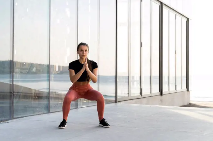 Young sportswoman doing squat exercise on promenade against glass window