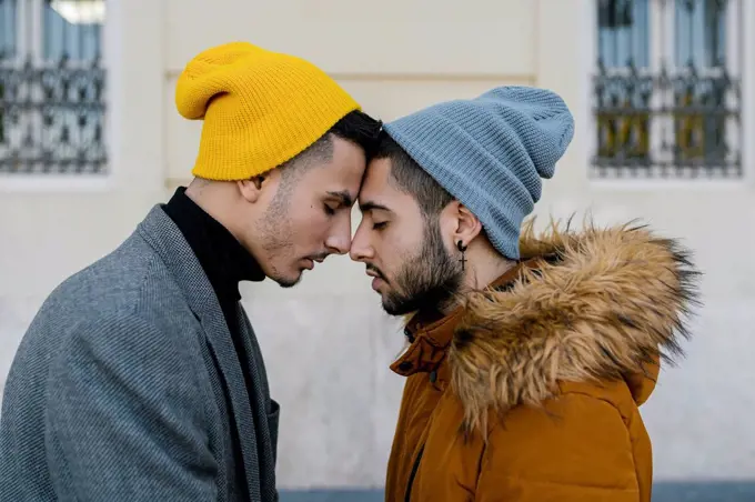 Gay couple wearing knit hat standing face to face with eyes closed against wall