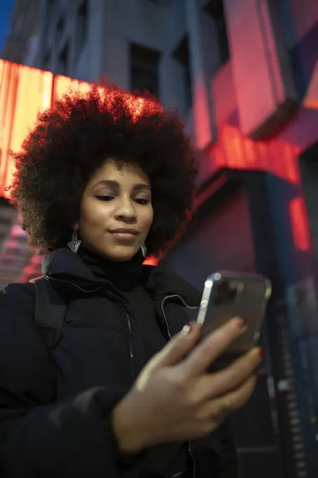 Afro young woman in warm clothing using smart phone in city at night