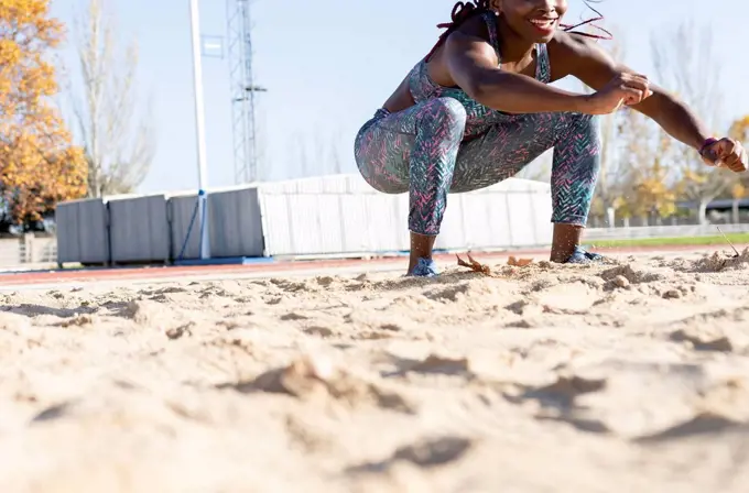 Smiling female athlete landing on sand in long jump during sunny day