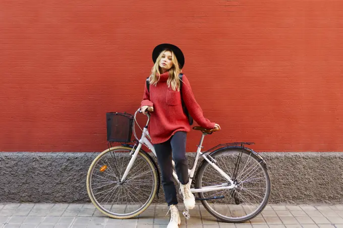 Young blond woman wearing hat standing with bicycle against red wall