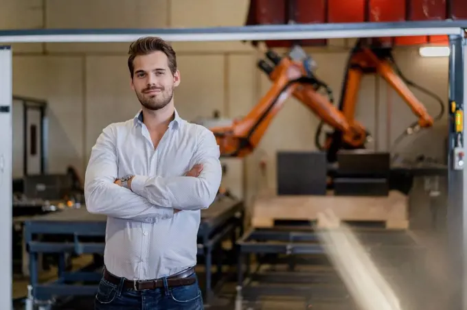 Young businessman standing with arms crossed against robotic arm at factory