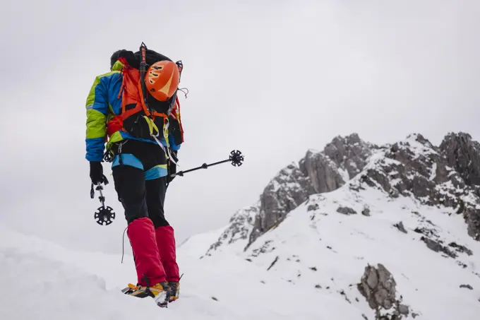 Mature man carrying backpack while walking on snowcapped mountain with crampon and hiking poles