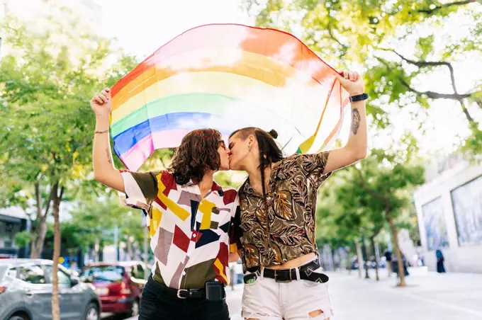Lesbian couple holding rainbow flag while kissing on footpath in city