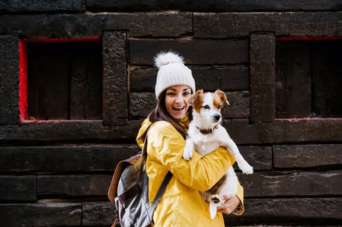 Portrait of smiling woman in yellow raincoat holding dog in front of log cabin