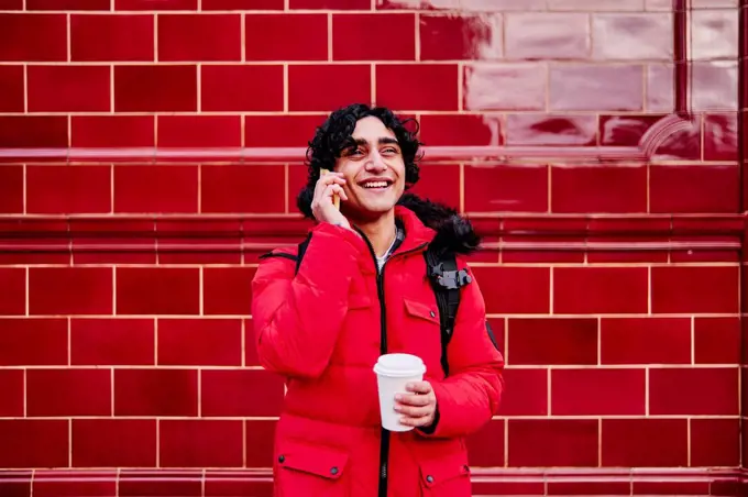 Smiling young man holding disposable coffee cup while talking on mobile phone against wall