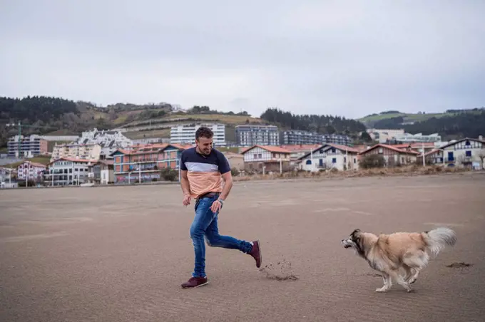 Man running with dog at beach near town against sky