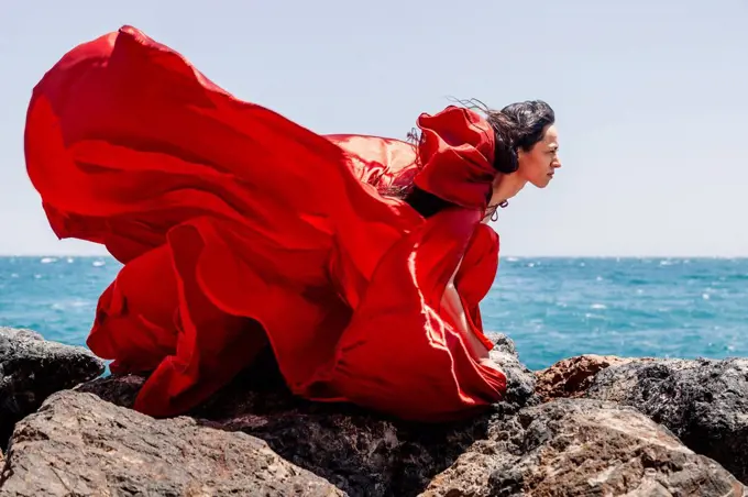 Confident female dancer in flowy red dress on rocks by seashore during sunny day