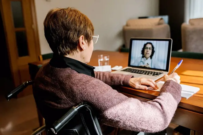 Senior woman sitting on wheelchair writing in book while video consultation at home during COVID-19