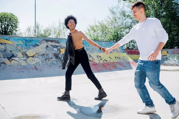 Playful young couple holding hands at skateboard park during sunny day